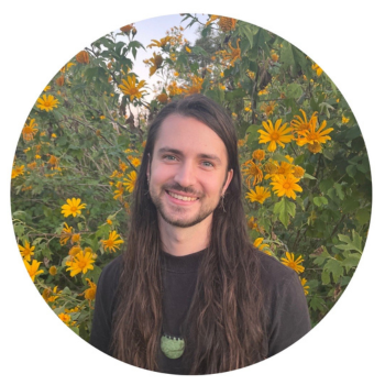 Smiling person with long hair standing in front of yellow flowers outdoors.